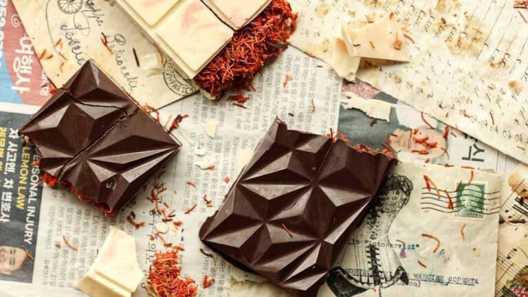 Close-up of geometric dark and white dubai chocolate bars with kataifi scattered around. The chocolates rest on vintage newspaper pieces, with a partially bitten piece showing texture.